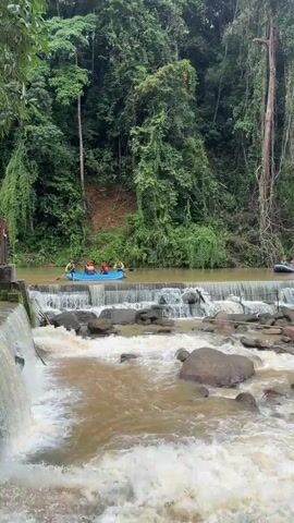 A group of people in two inflatable rafts are preparing to go down a small waterfall. The rafts are on a river, with dense green foliage and trees on either side. The water is murky brown, and there are rocks in the riverbed. The people in the rafts are wearing life jackets and appear to be ready for the descent. One raft has four people in it, and the other has three. They are all facing the waterfall.