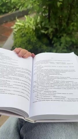This video shows a person's hand holding an open book outdoors. The book is white with black text and appears to be a novel. The person is wearing jeans. The background is blurry and shows green foliage and a brick walkway. In the later part of the video, the book is closed and the cover is visible. The cover is white with a colorful illustration and the title "Гимназия № 13". The person is wearing a pink watch on their left wrist. The video is shot from a first-person perspective.
