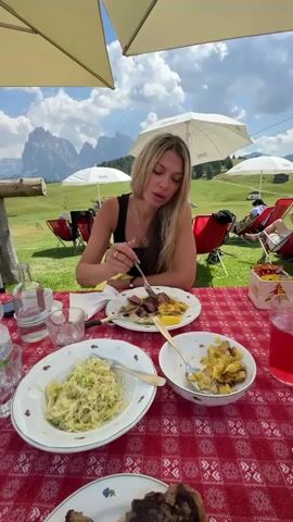 A blonde woman sits at a table with a red and white checkered tablecloth, eating a meal outdoors. She is wearing a black tank top and a yellow skirt, and has on a necklace and rings. In front of her are plates of food, including steak, cabbage, and potatoes, as well as drinks. Behind her, in the background, are mountains, green grass, and red chairs. The woman is actively eating, bringing food to her mouth with a fork.