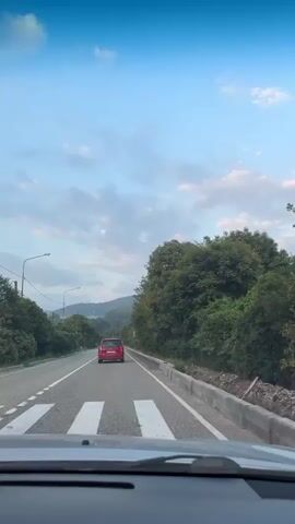 The video shows a view from inside a car driving on a two-lane road. A red car is driving ahead in the same direction. The road is bordered by trees and hills in the background. The sky is blue with some clouds. There are streetlights and power lines visible along the side of the road. The timestamp at the beginning of the video indicates the car is 600 meters away from a turn to the right.