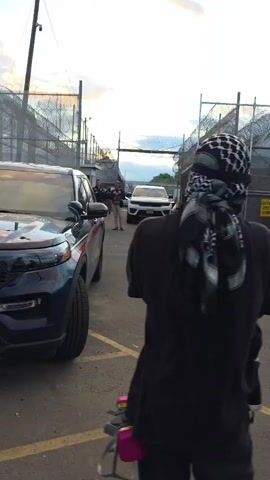 The video depicts a group of people wearing masks and bandanas, gathered outside what appears to be a correctional facility, recognizable by its high fences and barbed wire. Two police officers stand between two vehicles. One of the individuals carries a shield. The sky is light with clouds and what appears to be the sun setting.
