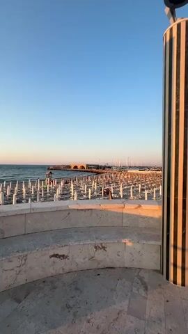 The video shows a beach scene at sunset. In the foreground, there is a curved stone structure with steps. To the right, a tall, striped pillar stands. Beyond the foreground, rows of white beach chairs and umbrellas are set up on a sandy beach. The sea is visible in the background, with a breakwater and a marina with boats. The sky is clear and blue, with a warm glow near the horizon. Several people are scattered across the beach, some sitting in chairs and others walking. The overall impression is a peaceful and scenic evening at the seaside.
