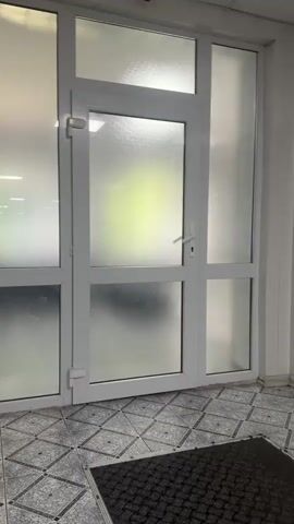 The video shows a white framed glass door with frosted panes. The door is positioned in a tiled entryway with a black doormat on the floor. The door is closed. Through the frosted glass, a blurry green and yellow light can be seen. The surrounding wall is white. The lighting is bright. The camera is stationary.