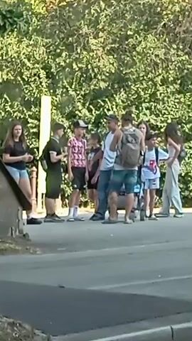 A group of ten individuals, appearing to be a mix of young adults and teenagers, stand on the side of a paved road. They are positioned near a tree line with lush green foliage. The individuals are casually dressed, with some carrying backpacks. One individual is holding a phone. The scene appears to be outdoors during daylight hours.