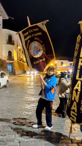 Here's a detailed description of the video:

The video appears to have been taken at night in an outdoor public space that seems to be a plaza or square, judging by the cobblestone ground and surrounding buildings with visible lights.

The main focus is on two banners that are being held by people. The first banner is dark with gold lettering. The words "CENTRO CULTURAL" are visible at the top, and there are other words below along with a circular image. The second banner, which is taller and more prominent in the frame, is also dark with gold lettering that reads "PERUANAS." It has an ornate gold trim.

In the background, several people are visible. A group of women are wearing traditional clothing with hats and colorful shawls. Other people are dressed in casual clothes, including jackets and jeans. There are tents or stalls set up in the background, suggesting some sort of event or market. Car headlights are also visible. 

The people holding the banners appear to be part of an organization or group that is involved in promoting Peruvian culture, as indicated by the banners' text. The group of women in traditional clothing suggests they may be participants in a cultural celebration or performance.