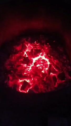 The video displays a close-up, overhead view of glowing red embers in a dark setting. The embers are clustered together, appearing as a pile of hot coals. The intense red and white light emanates from the center of the pile, suggesting significant heat. The surrounding area is obscured by shadow. There are no people or discernible objects other than the embers. The action, if any, is limited to the subtle flickering and shifting of the glowing coals. The environment is dark and appears to be contained, possibly within a fire pit or furnace.