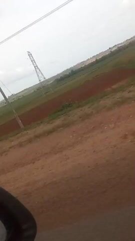 This is a low-resolution, shaky video captured from a moving vehicle, likely during daytime. The scene is an outdoor landscape with a dirt road in the foreground. In the middle ground, there are cultivated fields with some areas of brown soil and others with green vegetation. A tall, lattice-like electrical transmission tower is visible in the distance, along with overhead power lines. The sky is overcast. There are no people or animals visible in the frame. The overall impression is of a rural or agricultural area.