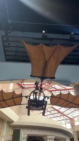 The video shows a model of an ornithopter, a flying machine that flaps its wings, suspended from the ceiling. The ornithopter is made of wood and fabric, with a wooden seat for the pilot. The wings are spread wide, and the entire structure is illuminated by natural light and artificial lighting. The background consists of a blue wall and a ceiling structure with red supports, suggesting an indoor environment like a museum or exhibition hall. The camera angle is looking upwards at the ornithopter.