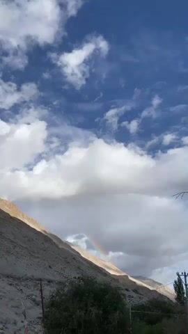A rainbow arcs across a partly cloudy sky above a dry, rocky mountainside. The sun illuminates a portion of the mountain, casting shadows on the rest.