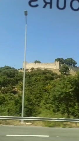 The video shows a stone fortress on a hilltop, surrounded by lush green trees. The sky is clear and blue. A road with a white stripe runs along the bottom of the frame. A lamppost is visible on the right side of the frame. The video appears to be filmed from a moving vehicle, as evidenced by the slight blur of the trees and the passing scenery. The fortress looks ancient, with battlements and weathered stone walls. The overall impression is of a historic site in a natural, picturesque setting.