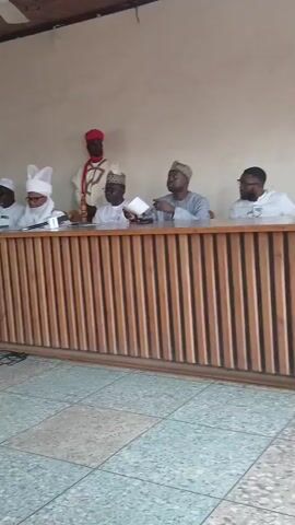 A group of men are seated behind a wooden desk in a room. A man in a red fez and robe stands behind them. Another man in a white robe is standing to the left of the desk, holding a microphone. A person holding a camera is standing in front of the desk, filming the scene. The room has a tiled floor and a drop ceiling with a fan.