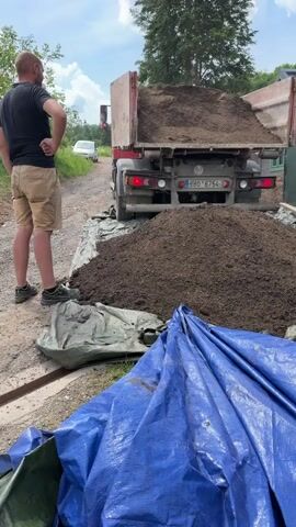 In a daytime outdoor scene in Nový Jáchymov, Czechia, a man stands on the left, observing a large truck as it dumps a significant pile of dark, earthy material, likely soil or compost. The truck's tipper bed is raised, releasing the material onto the ground, which is partially covered by a gray or green tarp. A bright blue tarp is folded in the foreground, next to the growing mound.

The man, dressed in a black t-shirt and tan shorts, with dark sneakers, stands with his hands on his hips initially, then briefly gestures with his right hand towards the truck. The truck is positioned on what appears to be a dirt or gravel path. Its rear features brake lights, a license plate reading "9BO 6754," and a small "80" speed limit sign. In the background, a white car is visible on the path, and lush green trees line the horizon under a partly cloudy sky. The overall impression is an active worksite or delivery of bulk material.
