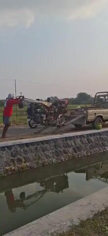 In an outdoor scene, a person wearing a red shirt and dark shorts is pushing a two-wheeled tractor up a ramp towards the bed of a light-colored pickup truck. Another person, partially obscured, appears to be operating a motorcycle that is positioned next to the tractor on the ramp. The truck is parked alongside a stone-lined canal, which contains water. The ground is dirt, and in the background, there are fields and trees under a bright sky. The overall impression is one of agricultural activity or equipment transport.