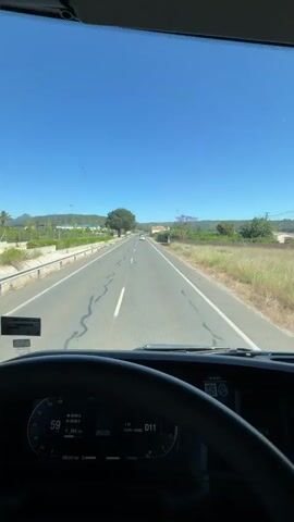 A view from inside a truck as it drives along a straight road on a sunny day. The road has white dividing lines and a small concrete barrier on the left side. Vegetation grows on both sides of the road. In the distance, there are houses and mountains. The truck's dashboard is visible, displaying information like speed and mileage. Music is audible in the background.