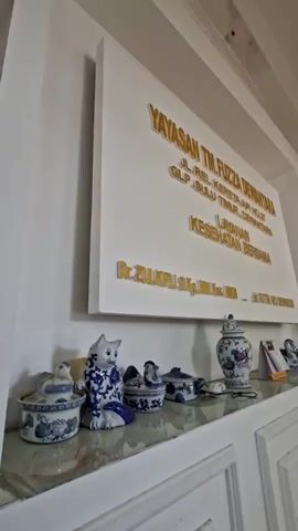 The video shows a wall with a sign that reads "YAYASAN TM. FUZZA DEWANTARA JL. REL. KERETA API NO.27 GLP. SULU TIMUR. DEWANTARA LAYANAN KESEHATAN BERSAMA Dr. ZULKIFLI S.Kp., M.Kes., LHKKN" (Foundation of TM. Fuzza Dewantara, Health Services Together).  Below the sign is a shelf with several blue and white porcelain figurines, including a cat, chickens, and a vase. The wall and shelf are white. The floor is tiled. The video pans slightly to the right, revealing a doorway into another room.