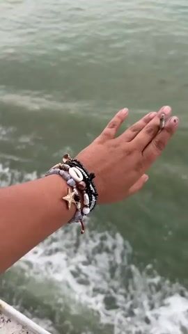 A close-up shot shows a person's arm adorned with multiple bracelets against a backdrop of turbulent green-blue water. The bracelets are of various styles, including beads, shells, and a starfish charm. The person's hand holds a lit cigarette between the fingers. The arm extends slightly over what appears to be a white railing, suggesting the person is on a boat or pier. The water displays foamy white caps, indicating windy conditions.