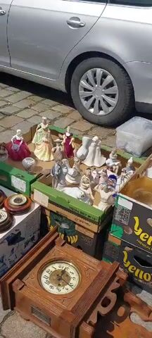 The video shows a variety of antique items laid out on tables and boxes, likely at an outdoor market. In the foreground, a wooden clock with a visible pendulum and Roman numerals is on display. To the left of the clock, several circular wooden objects, possibly barometers or clocks, are stacked. Behind these items, a blue tarp covers a collection of objects including porcelain figurines of people in vintage clothing, decorative lamps with colorful glass shades, and a small white bell. Further back, boxes contain more figurines, including some that appear to be porcelain. A car is partially visible in the background, parked on a cobblestone surface. The overall impression is of a bustling antique stall with a wide assortment of decorative and functional items for sale.