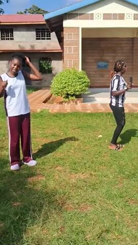 Several individuals are dancing outdoors on a grassy lawn. The setting appears to be a residential or communal area, with buildings visible in the background. People are dressed in casual clothing. The scene includes elements like trees, a car, and architectural structures. The video shows them moving and dancing in a seemingly joyful manner.