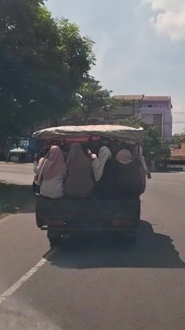 A video captured from a moving vehicle shows the rear of a small, open-air transport vehicle filled with passengers. The passengers, predominantly women wearing headscarves, are seated closely together in the back of the vehicle, which appears to be a modified utility vehicle or cart. The vehicle is driving on a paved road under a bright, sunny sky with scattered clouds. Trees line the side of the road, and buildings are visible in the background. The overall impression is of a form of local transportation in a potentially rural or semi-urban setting.
