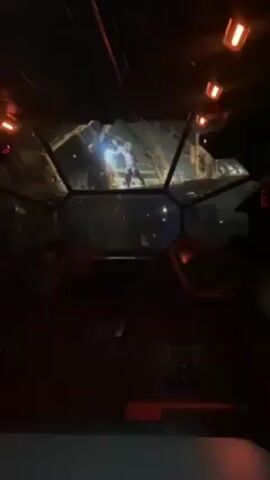 The video presents a first-person perspective from inside a cockpit. Through the hexagonal windshield, an animated battle scene is visible, featuring what appears to be a figure in combat. The cockpit interior is dark, illuminated by red lights on the control panels and overhead. The scene suggests a simulation or ride experience, possibly themed around science fiction or space.