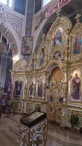 The video shows a view inside a church, focusing on an ornate, golden iconostasis. The iconostasis is decorated with intricate carvings and houses numerous religious icons depicting figures in traditional attire. The icons are set within arched frames, and the entire structure is gilded, reflecting light. To the left, a portion of a staircase is visible, leading up to the iconostasis. A small altar or stand with an icon is placed in the foreground. The walls of the church are decorated with religious paintings and murals, including text in a foreign language. The overall impression is one of a richly adorned religious sanctuary.