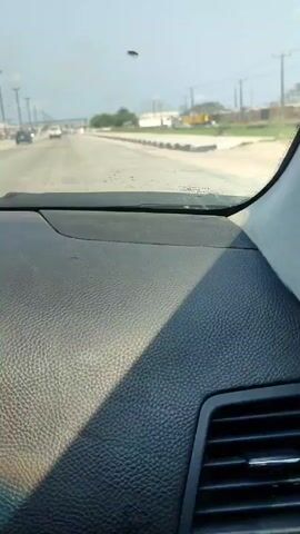 The video shows a view from inside a car driving on a highway. The dashboard is visible in the foreground, with a textured black surface. A strong shadow crosses diagonally across the dashboard. The road ahead is wide, with multiple lanes separated by concrete dividers.  There are streetlights lining both sides of the highway.  Other vehicles are visible on the road, including cars and a white truck. Buildings and greenery are seen in the background. The sky is bright and slightly overcast.  Music is playing in the background.