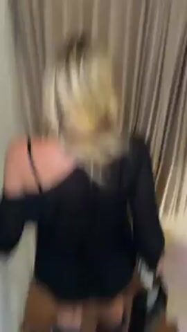 A blonde woman walks down a flight of wooden stairs. She is wearing a black, one-shoulder dress and black boots. She is carrying a black bag in her right hand. The stairs are next to a beige curtain, and a window is visible at the bottom of the stairs.