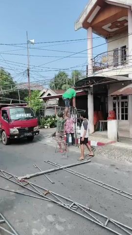 In a residential street, four men are working on dismantling a metal truss structure that appears to be supporting an overhead canopy. A red truck is parked on the left side of the street. Two men are standing on a ladder-like structure, one of whom is wearing a green shirt and is positioned higher up, seemingly disconnecting pieces of the truss. Another man in a pink shirt is assisting from below, holding onto the structure. A fourth man in a white and black shirt is standing nearby, looking on. On the right side of the street, a building with a covered porch is visible. Several long pieces of the metal truss are lying on the asphalt road. The sky is clear and blue.