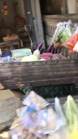 The video opens with a blurred close-up of a wooden cart overflowing with various fresh produce. Visible items include eggplants, cucumbers, carrots, and what appear to be gourds or squash. The cart is positioned outdoors, with hints of a shop or market stall in the background. In the foreground, several plastic bags containing food items, possibly snacks or packaged goods, are also blurred. The overall impression is of a bustling, outdoor food vendor. The camera movement is unsteady, suggesting it's handheld. No people are clearly discernible in the frame.