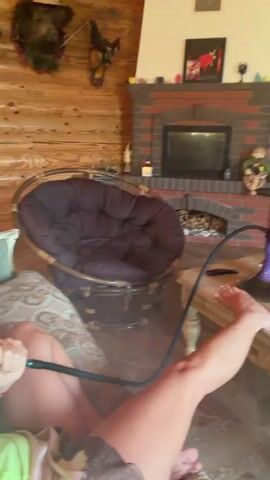 A person is sitting on a couch, holding a hookah hose and inhaling smoke. The person is wearing a yellow shirt and appears to be in a living room with a fireplace and a large armchair. The camera zooms in on the person's face as they exhale smoke.