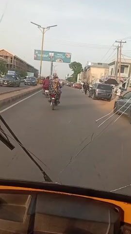 The video shows a view from inside a yellow, three-wheeled vehicle moving along a paved street. The street is relatively straight with several lanes of traffic. There are motorcycles, cars, and a similar three-wheeled vehicle visible on the road. Pedestrians are walking along the sidewalks on either side of the street. Buildings line both sides of the street. Above the road, a large billboard advertises Dangote Salt. The sky is clear and light.