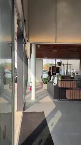 The camera slowly pans across the interior of a Porsche dealership. The sliding glass doors open to reveal a modern showroom with gray tile floors. On the left, a white Porsche sports car with a red interior is displayed next to a black tablet stand. On the right is a light silver Porsche SUV. A man wearing jeans and a black shirt walks past the SUV. The walls are painted white and the ceiling is partially covered with a dark brown patterned wood design. The word "PORSCHE" is written in large letters on a black background along the back wall. A quote is written on the wall next to the white sports car.