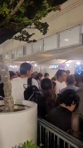 A crowd of people are gathered outdoors at night under large umbrellas. The people are of various ages and genders and appear to be in a line or milling about. Some individuals are looking at their phones. In the background, there are lights and signs, including one that reads "CONEY IS". The area appears to be an outdoor dining or entertainment venue.