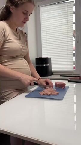 A pregnant woman in a beige dress is standing in a kitchen and chopping chicken on a blue cutting board. To her right, there is a piece of raw red meat. Behind her, there is a window with white blinds and a black appliance. The counter is white.
