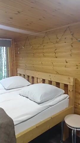 The video shows a bedroom with wooden walls and ceiling. Two beds with white linens and pillows are visible. On the left bed, a brown blanket is draped, with folded white towels placed on top. A large window looks out onto a forest of birch trees. The room is lit by a square ceiling light and a string of small lights along the wall. A round stool is partially visible in the lower right corner.