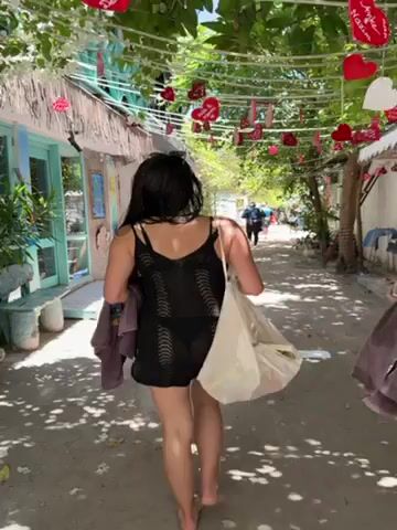 A woman with dark hair, wearing a black spaghetti-strap dress, walks away from the camera. She carries a light-colored tote bag over her shoulder and a dark-colored garment in her left hand. Another person, presumably a young woman or girl with long dark hair and wearing a white top and skirt, walks beside her. The scene takes place on a sandy path with various buildings on either side. The buildings are painted in light colors, including white and turquoise, with some having thatched roofs. Overhead, a string of red heart-shaped decorations hangs between the buildings. Trees are visible, providing shade along the path. The overall environment suggests a tropical or beachside location.
