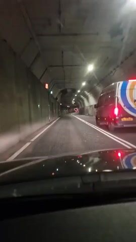 The video captures a first-person perspective from inside a vehicle driving through a dimly lit road tunnel in North Tyneside, United Kingdom. The time appears to be night or twilight, as the tunnel relies entirely on artificial lighting.

The vehicle is proceeding slowly, following a white van with red taillights illuminated. The van has a large circular logo on its rear, featuring blue, orange, and white colors, with the letters "CT" prominently displayed. Below the logo, a phone number and website address are visible but illegible. The van's license plate reads "GL21 AND."

The tunnel's walls are made of concrete or similar material, appearing grey and textured. On the left side, there are several green doors set into the wall, possibly for emergency exits or access to maintenance areas. Overhead, numerous light fixtures illuminate the roadway, and at one point, two large ventilation fans are visible mounted on the ceiling. The road is asphalt, marked with solid white lines separating the lanes and a dashed white line on the right shoulder. Reflections of the vehicle's dashboard and the van's taillights are visible on the windshield at the bottom of the frame. The tunnel appears to have a slight curve to the right in the distance.