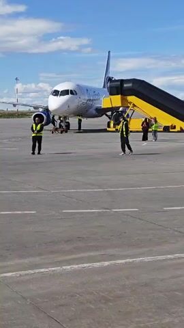 The video shows an airplane parked on an airport tarmac under a partially cloudy blue sky. A set of yellow and black airstairs is connected to the airplane's door. Several individuals in reflective vests are visible on the tarmac near the airplane, appearing to be airport personnel. One person in a vest walks from left to right in the foreground. Two other individuals in vests stand near the airstairs. Two more individuals, one in red and one in green, stand at the top of the airstairs. The scene is static, with no discernable movement of the airplane or significant activity.