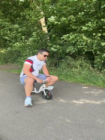 A man is sitting on a small white bicycle on a paved path. He is wearing a white t-shirt with red and blue accents on the sleeves, grey shorts, and blue and white sneakers. He has sunglasses on and is looking down at the handlebars of the bicycle. The bicycle has black tires with colorful spokes. The path is surrounded by green trees and grass. The man is holding the handlebars and his feet are on the ground. He appears to be moving the bicycle slightly forward and backward.