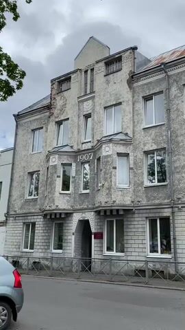 A street-level view of a weathered, multi-story building with ornate detailing and the year 1907 inscribed above the central arched entrance. The building's facade is predominantly gray, with sections of lighter brickwork on the lower level and a rough stucco finish on the upper floors. Numerous windows of various sizes are visible, many with white frames. The roofline features decorative elements and appears to have some signs of age, including rusted metal sheeting. A few trees and a cloudy sky form the background. A small section of a gray car is visible in the lower left corner of the frame.