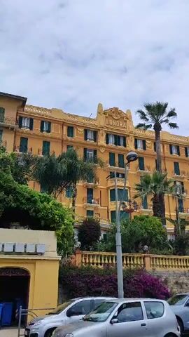 The video shows a building called "Londra" under a cloudy sky. The building is painted in a light yellow color and has multiple stories. Green shutters adorn the windows of the building. A couple of palm trees are planted next to the building. In front of the building, there is some landscaping with purple flowers and a stone wall. There is a street with several parked cars below.