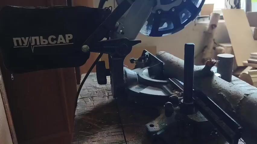 The video shows a miter saw cutting a piece of wood. The saw is labeled with "ПУЛЬСАР". Sawdust is flying from the cut. The saw is on a wooden table in a workshop or garage. There are other pieces of wood in the background.