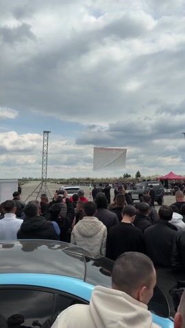 A crowd of people watches two cars engage in a drag race at an outdoor event, with many recording the event with their phones. The race takes place on a paved surface under a cloudy sky with a large banner in the background. One of the vehicles has "RTG GARAGE" labeled on its side, kicking up a cloud of dust as it accelerates.