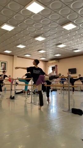 The video shows a ballet class in session. Several young people are practicing ballet moves at a barre in a dance studio. The studio has a polished wooden floor and a tiled ceiling with fluorescent lights. A portrait of a man in a suit hangs on the wall. One young woman in the foreground is demonstrating a ballet pose, with her arms extended and one leg lifted. Other students are positioned along the barre, some in similar poses, while others are sitting on the floor. The atmosphere appears to be one of focused practice and instruction.