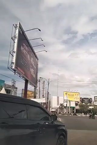 The video shows a street scene with several billboards. In the foreground, a black SUV is driving on the right side of the road. In the background, there are several buildings and more billboards. One billboard, prominently featured on the left side of the screen, is large and displays text in red and white. Another billboard in the distance is yellow and features a picture of a man in a suit along with other individuals. The sky is overcast with clouds. A motorized tricycle carrying a large load of white material is also visible on the road. The overall impression is a commercial area with advertising being a significant visual element.
