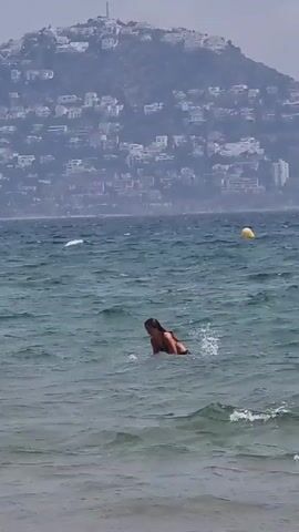 A person is swimming in the ocean. The water is choppy, with waves breaking on the shore in the foreground. In the distance, a hill is covered with houses and trees. A yellow buoy is visible in the water. The person is in the middle of the frame, facing left. They are wearing a swimsuit and appear to be reaching for something in the water.