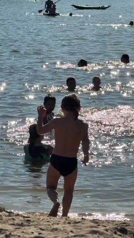 The video shows a sunny day at a lake or beach with several people in the water. In the foreground, a child wearing black swim trunks splashes water as they walk towards the camera. Behind them, other children and adults are wading and swimming. In the background, a person is paddleboarding and another is on a kayak. The water is shimmering with sunlight, and there is a sandy shoreline.