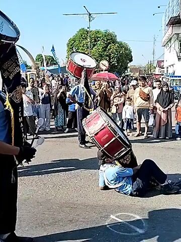 A medium shot on a sunny day captures a street parade with performers and onlookers. In the foreground, a person in a blue uniform and black gloves stands facing right, holding a large bass drum on their head with one hand, while holding a drumstick in the other hand. Behind them, another person in a blue uniform is lying on their back on the street, balancing another large bass drum on their chest and feet. Further down the street, a third person in a blue uniform is also balancing a smaller red drum on their head while standing. A large crowd of people, some holding umbrellas, watches the parade from the sidelines. The street is paved, and there are buildings and trees in the background. The drums are red and white.