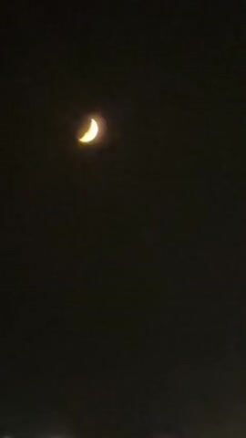 A crescent moon hangs in a dark night sky over a body of water. The moon is illuminated and appears to be a pale yellow color. In the distance, along the horizon, there are faint lights that suggest a city or town. The water in the foreground has small waves breaking on a dark shore. The overall scene is dark and tranquil, with the moon and distant lights providing the only significant illumination.