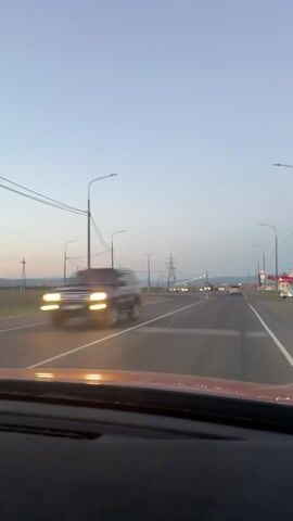 The video shows a first-person view from inside a car driving on a road at dusk. The sky is a pale blue with hints of pink and orange on the horizon, suggesting the sun has recently set or is about to rise. A highway with multiple lanes stretches ahead, lined with streetlights and utility poles carrying power lines. In the distance, there are a few other vehicles on the road, their headlights visible. A large, lattice-like structure, likely a power pylon, stands prominently in the middle ground. To the right of the road, a red and white building, possibly a gas station or convenience store, is visible. The road curves gently to the right, leading towards the distant lights. The car's dashboard is partially visible in the foreground. The overall impression is of traveling on a highway during twilight.